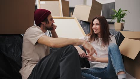 A-brunette-girl-in-a-white-T-shirt-communicates-with-her-boyfriend-in-a-red-hat-with-stubble-while-they-sit-near-a-sofa-covered-with-black-polyethylene-among-a-large-number-of-boxes-and-a-house-plant-in-a-modern