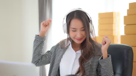 close up of a hard-working, young, healthy asian woman surrounded by packages ready to be shipped takes a music and dance break at her desk