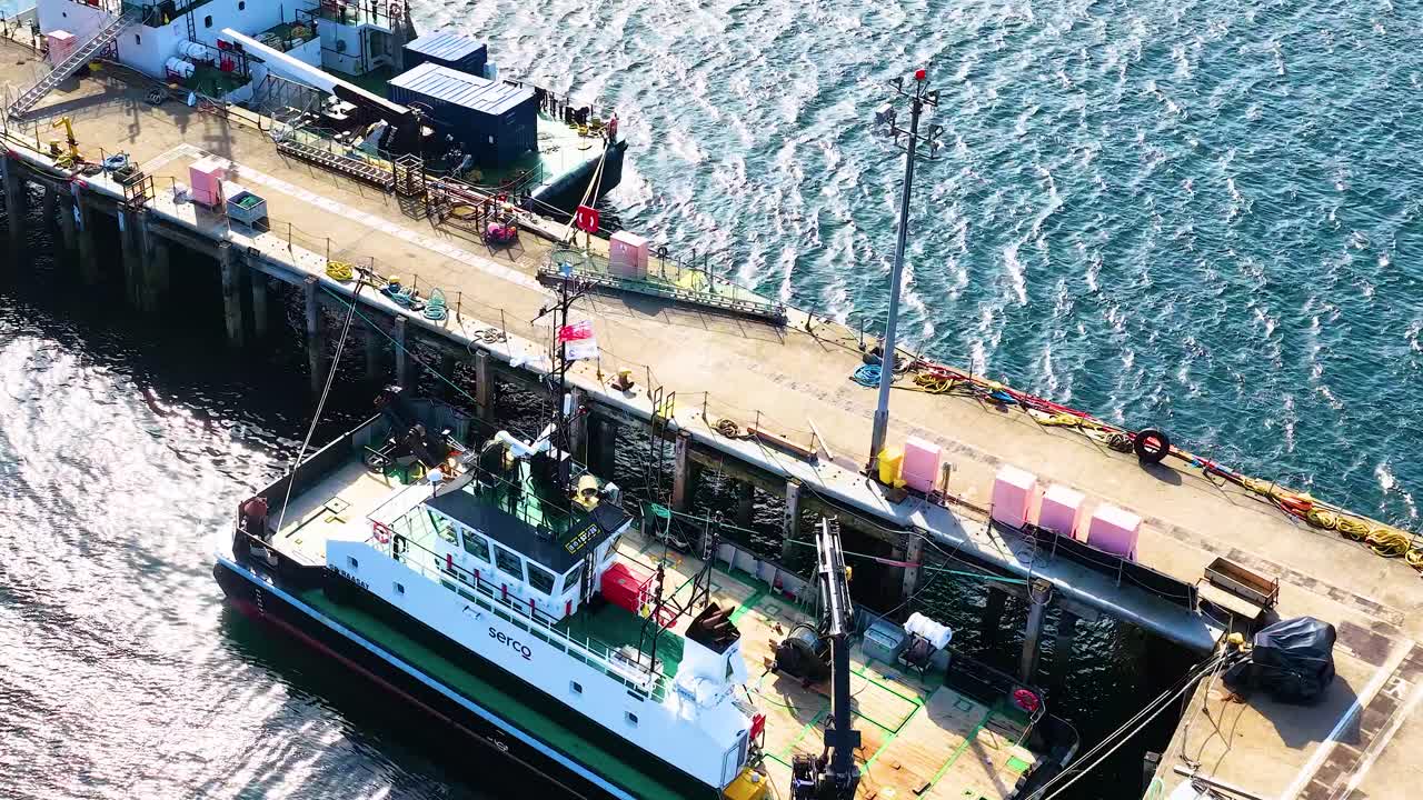Fishing vessel approaches and docks at sunlit pier in Dundee, Scotland, captured by aerial camera