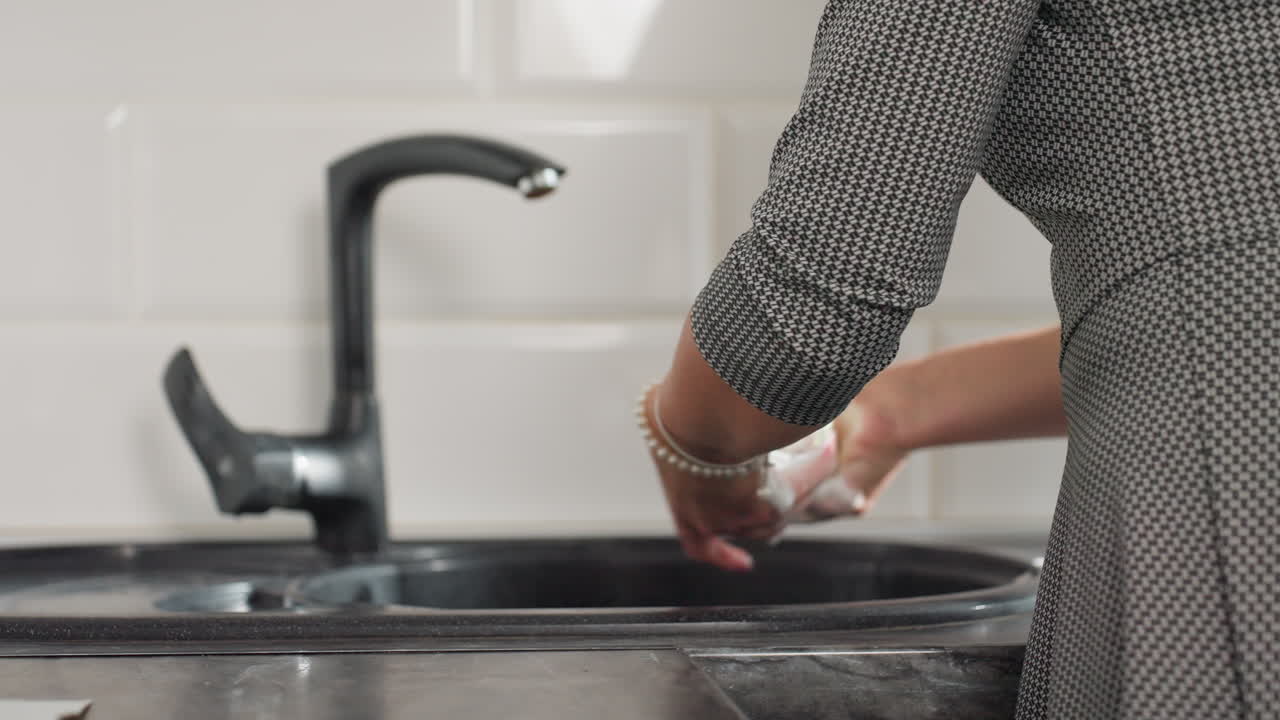 Woman thoroughly washing glass cup at kitchen sink wearing bracelet with focus on cleanliness hygiene and household chore routine showing careful attention to detail in modern bright kitchen