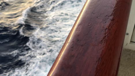 static view overlooking a wooden hand rail on a ship to see the waves breaking in the wake of the boat and sea foam with golden sunlight reflecting on the water