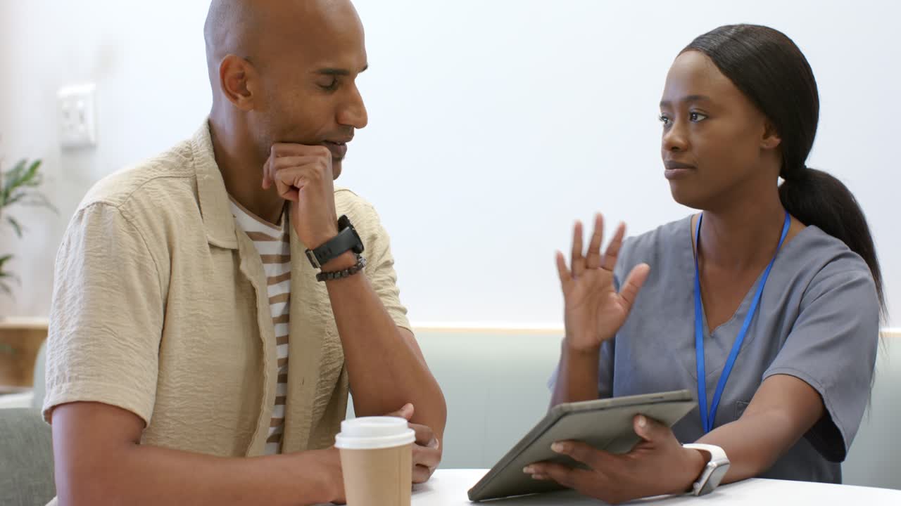 Woman in scrubs showing tablet, gesturing while explaining care in clinic; man listening with cup