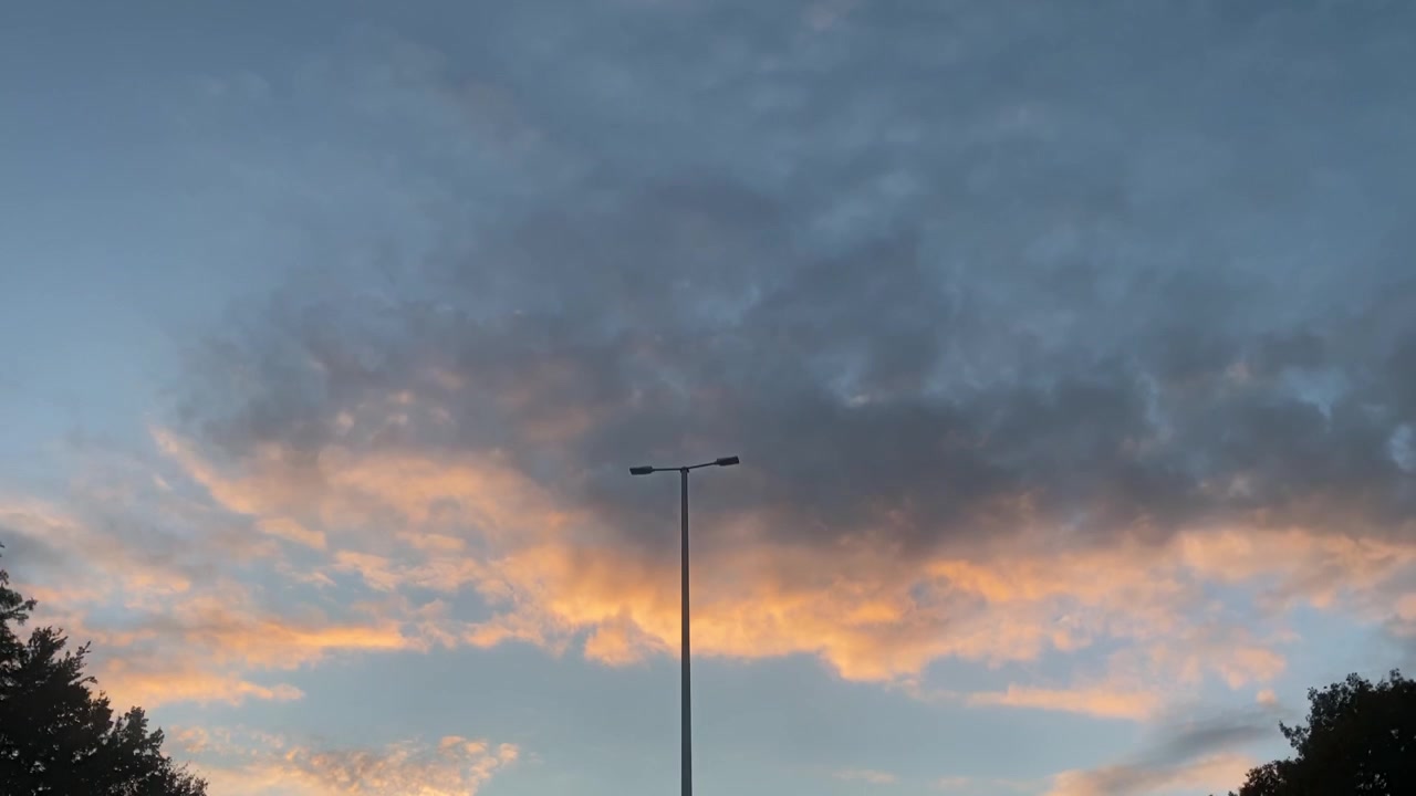 Awan senja yang membara di atas langit biru yang tenang dengan pepohonan dan tiang lampu di latar depan