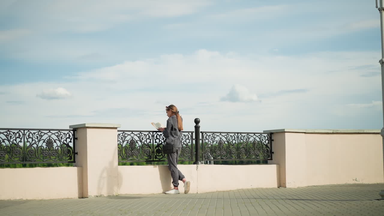 Lady in grey clothing and black handbag stands close to fence, resting her right knee while reading a book, trees visible in the distance and a lamp post nearby