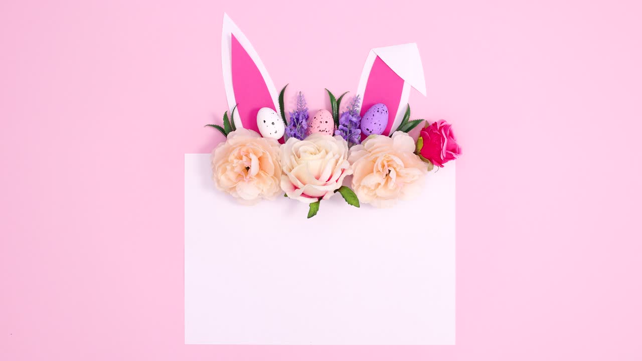 토끼 귀와 함께 부활절 꽃 장식은 파스텔 분홍색 배경에 종이 카드 메모와 함께 나타납니다.