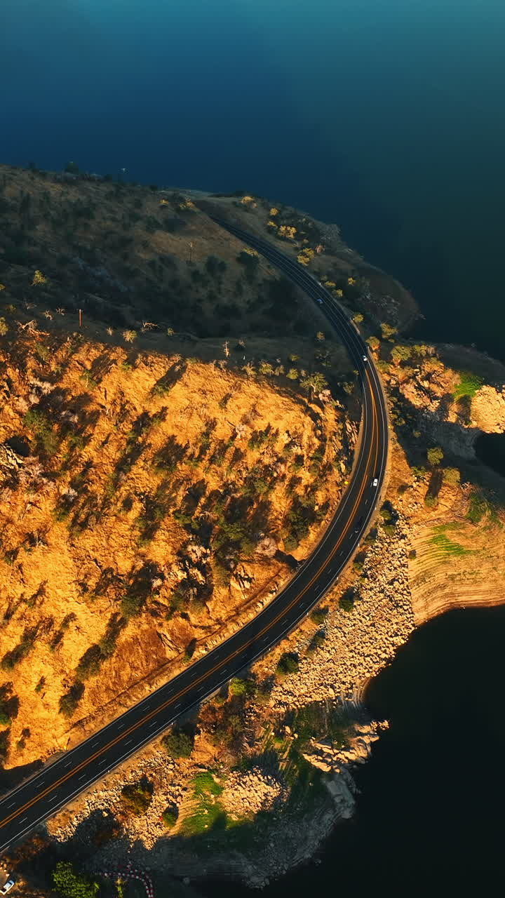 Speed highway circumflexes the mount with rarely growing trees. Beautiful calm lake is at the foot of the mountain. Top view. Vertical video