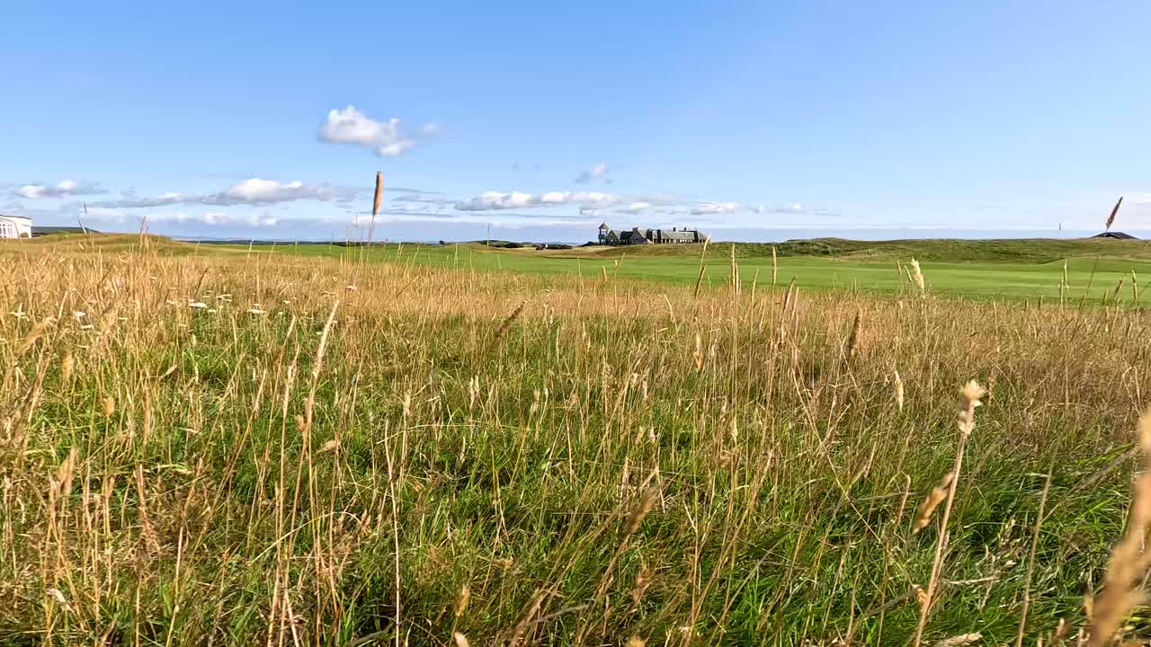 Camera slowly pans over tall grass, revealing golf course, hotel, blue sky, and summer landscape