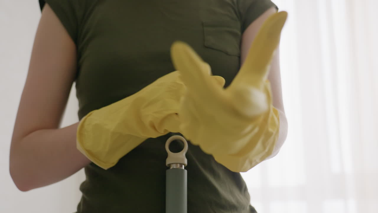 Close up of cleaner in yellow gloves adjusting glove by stretching fingers with mop handle resting on stomach while preparing for cleaning duty indoors against bright blurred background