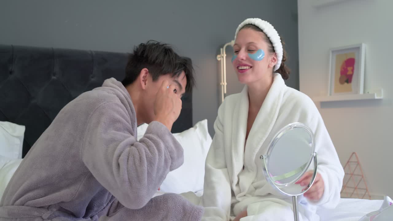 With blue patches Asian man and African woman in bathrobes holding mirror checking patch fit