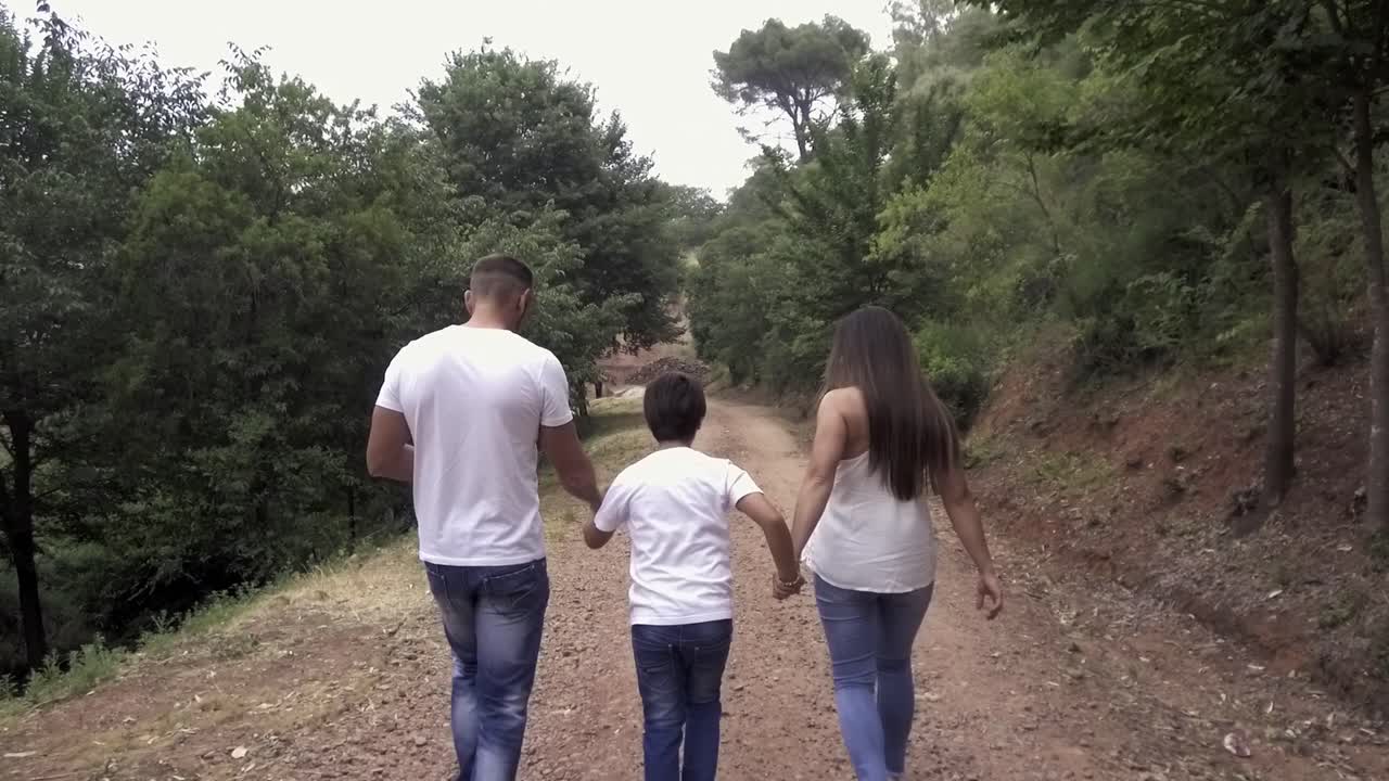 madre y padre caminando por un camino de tierra en la naturaleza, sosteniendo las manos de su hijo en el medio, todos vestidos de blanco y azul