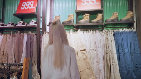 modern mother moves to jeans rack in clothing store, browsing denim pairs with gentle touch, fingers scanning textures and sizes while blurred shoppers move along aisles under warm lights