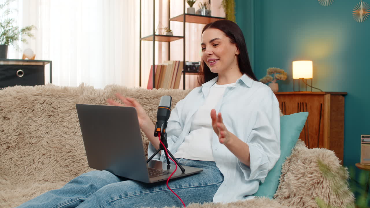 Young woman sitting on sofa recording podcast speaking seriously into microphone with focused look