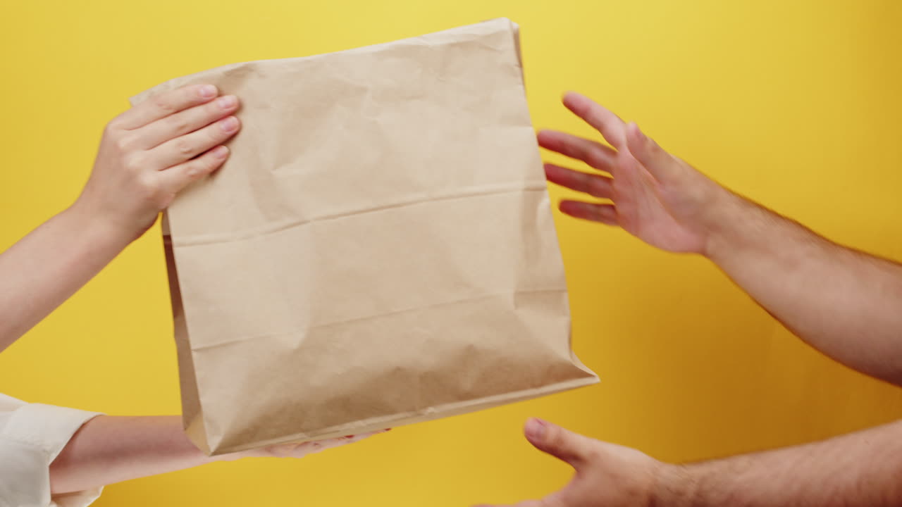 Hands Exchanging Brown Paper Bag Against Yellow Background
