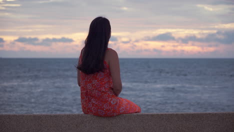 Happy-Korean-woman-in-30th-admiring-a-spectacular-sunset-sitting-on-seafront,-Stunning-cloudy-dramatic-sky-above-the-endless-seascape-in-slow-motion