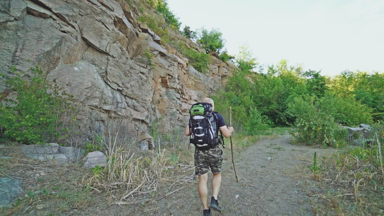 a traveler with back is walking on the path with a stick in his hand near the rocks in the summer day on the background of green trees
