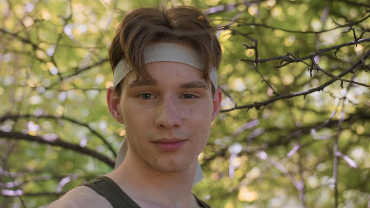 young man turns to camera with relaxed smile while insects hover around sweaty face, bandage tied on brow, sunlight filtering through branches, foliage rustling in calm thicket under tree