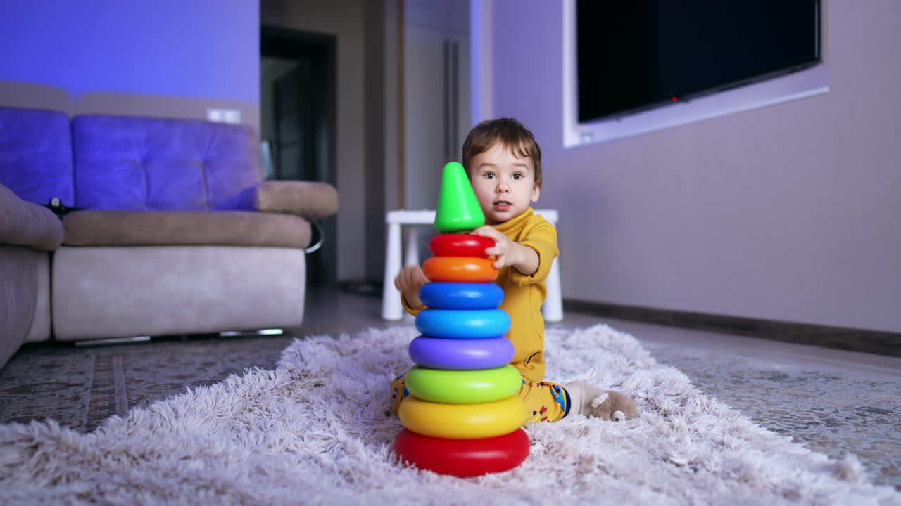 Cute baby in yellow pajama tries hard to disassemble toy pyramid. Lovely toddler boy playing at home.
