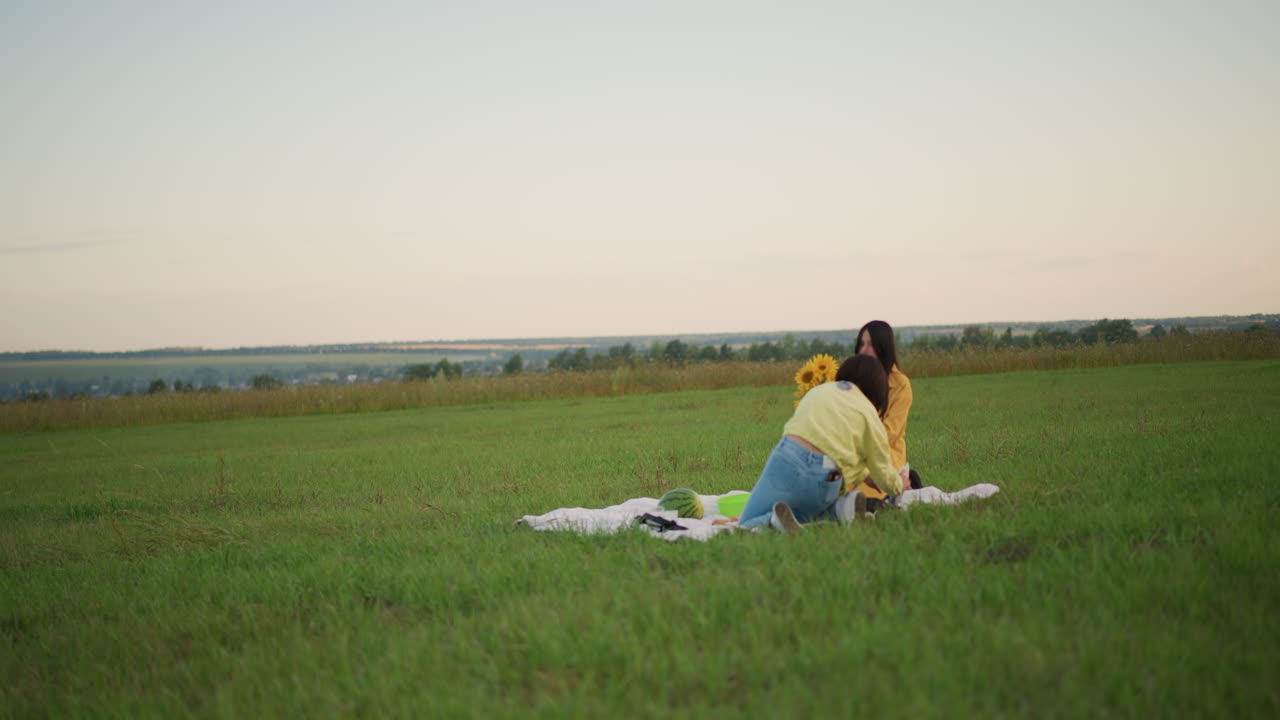 Two women on blanket in meadow sharing sunset conversation, yellow jackets and denim jeans, smiling and laughing, casual weekend picnic, candid friendship moment, pastoral skyline and soft dusk light