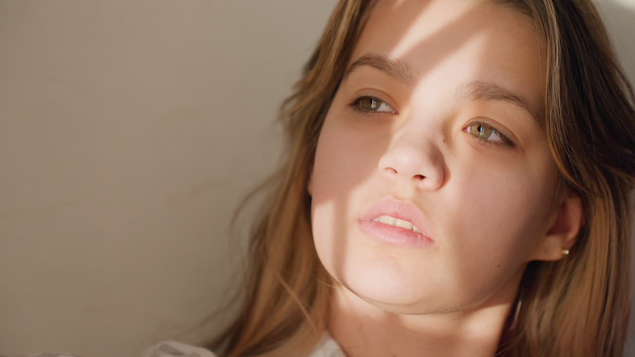 Ung, kaukasisk kvinde i blødt sollys, vinduesskygger hen over ansigtet, introspektivt blik, naturlig makeup, varme hudtoner, minimal indendørs opsætning, filmisk nærbillede, der fanger eftertænksom stemning