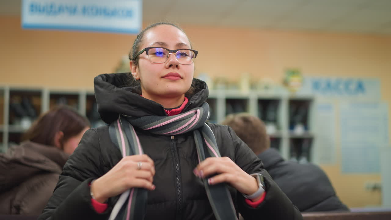 Damsel with glasses wearing black winter jacket and striped scarf adjusting hair indoors, candid moment in public waiting area with other people around, natural expression, relaxed atmosphere