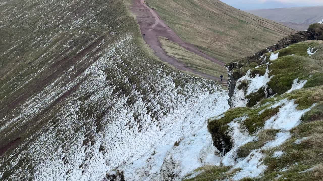 Stretta cresta innevata su Corn Du con un escursionista solitario che cammina sopra una valle ripida nei Brecon Beacons, creando un drammatico paesaggio montano con profondità e un'atmosfera avventurosa.