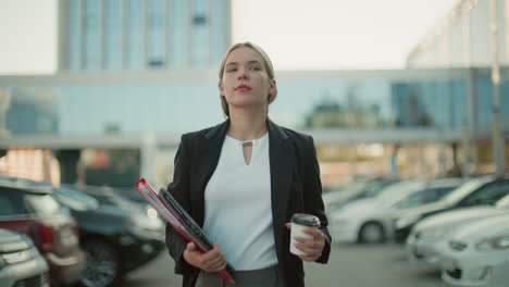 confident businesswoman walking through urban parking lot holding coffee cup and file folder with focused expression in modern city setting with office buildings and parked cars
