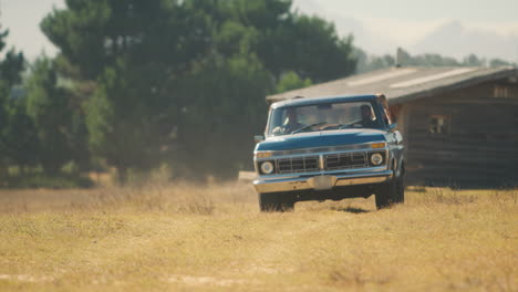 Friends-Driving-in-Pick-Up-Truck-Across-Field-On-Road-Trip-With-Cabin-In-Background