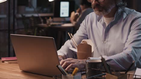 grey haired man with eyeglasses and blue shirt sitting in front of computer while eating food and working