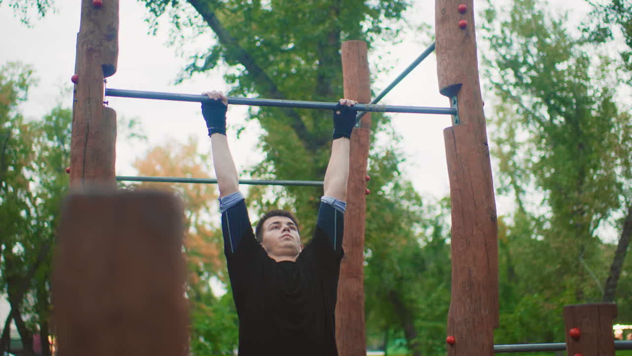 Pullups performed in park, Outdoor workout with intense arm effort, Man strains during pullups in leafy park setting, Solitary man engages in strenuous pullup exercise among trees in park