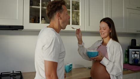 happy pregnant brunette woman feeding her husband fruit salad using a fork in the kitchen in the morning