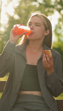 dama vestida de gris sentada en un banco del parque al aire libre, comiendo un bocadillo y sosteniendo una botella de bebidas de jugo de él, en un entorno de parque tranquilo, está rodeada de exuberante vegetación y árboles