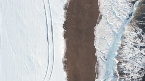 Still-aerial-above-waves-crashing-on-winter-Icelandic-beach-with-snow