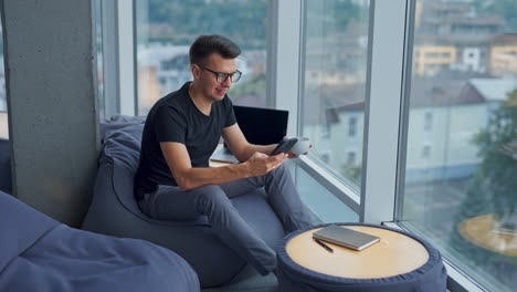 dark-haired man in glasses sitting on bean bag chair holding a phone and a cup. a break from work at computer. panoramic windows in blur at backdrop.