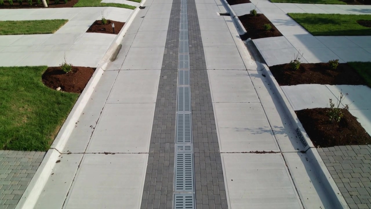 Telecamera dall'alto che documenta una griglia di scarico lineare su un marciapiede suburbano, valutando il flusso di drenaggio
