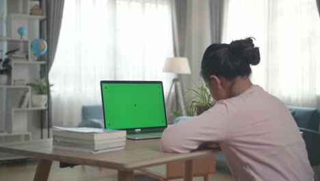teenage girl sitting at her desk learning online on a laptop with green mock-up screen in her living room and writing on book