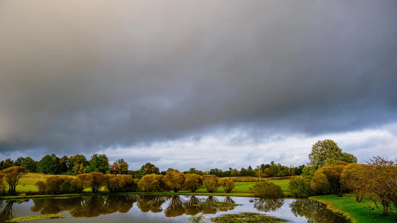 Vroege herfst timelapse op het platteland met een vijver op de voorgrond en dikke wolken die door de lucht bewegen. Gele en oranje bladeren aan de bomen.