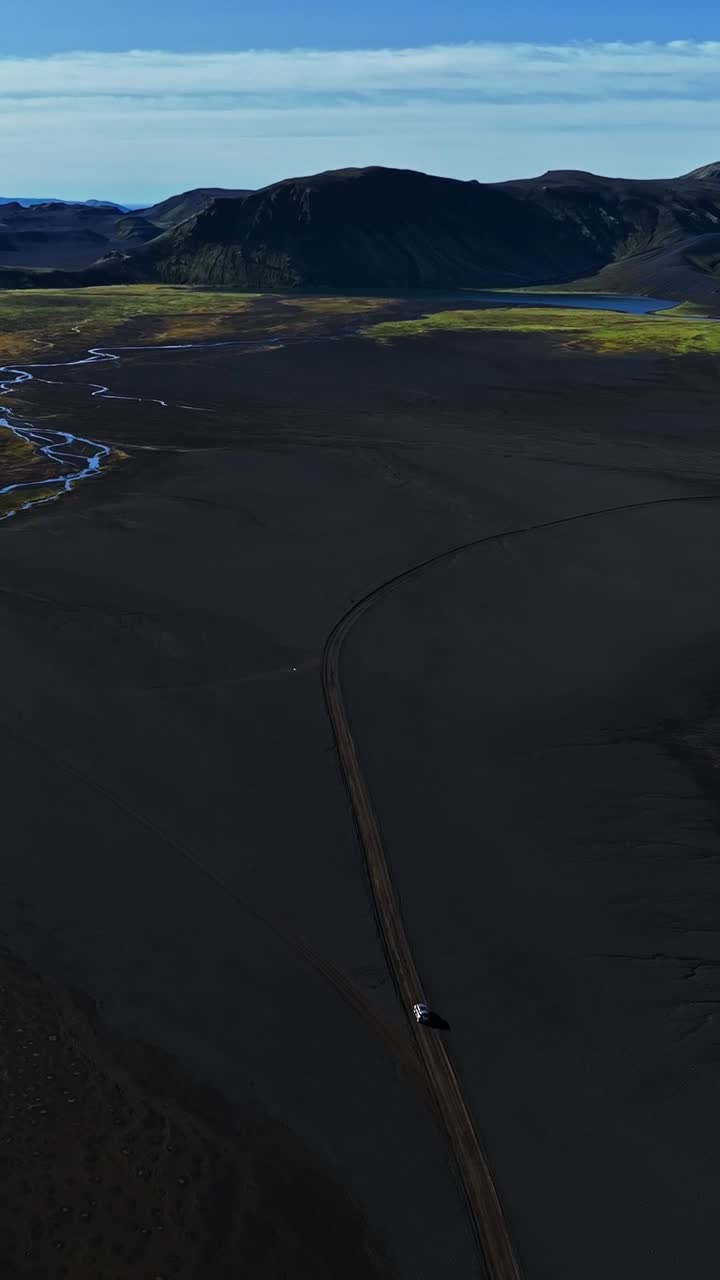 Ripresa aerea verticale di un SUV su un sentiero solitario attraverso la pianura vulcanica nera dell'Islanda, con montagne distanti, un lago e corsi d'acqua intrecciati che creano una drammatica scena di viaggio in luoghi remoti