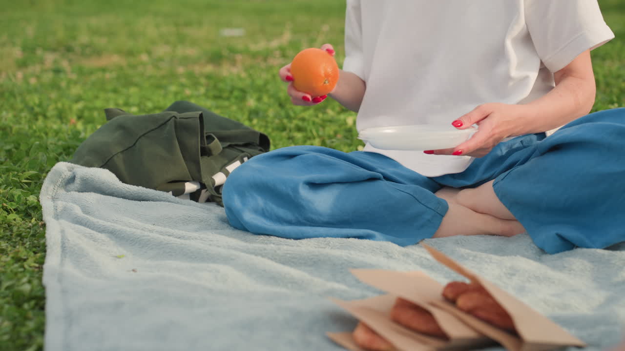 lower view of picnic woman taking out plate and orange from canvas bag on blanket, hands arranging healthy snack during relaxed outdoor summer outing on green grass under soft sunlight