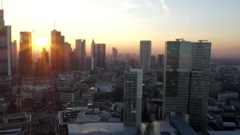 Bengalas-De-Sol-Que-Se-Revelan-Entre-Los-Rascacielos-De-Frankfurt-Am-Main,-El-Horizonte-De-Alemania-En-La-Hermosa-Luz-Del-Atardecer-De-La-Hora-Dorada,-El-Camión-Aéreo-Se-Desliza-Hacia-La-Izquierda