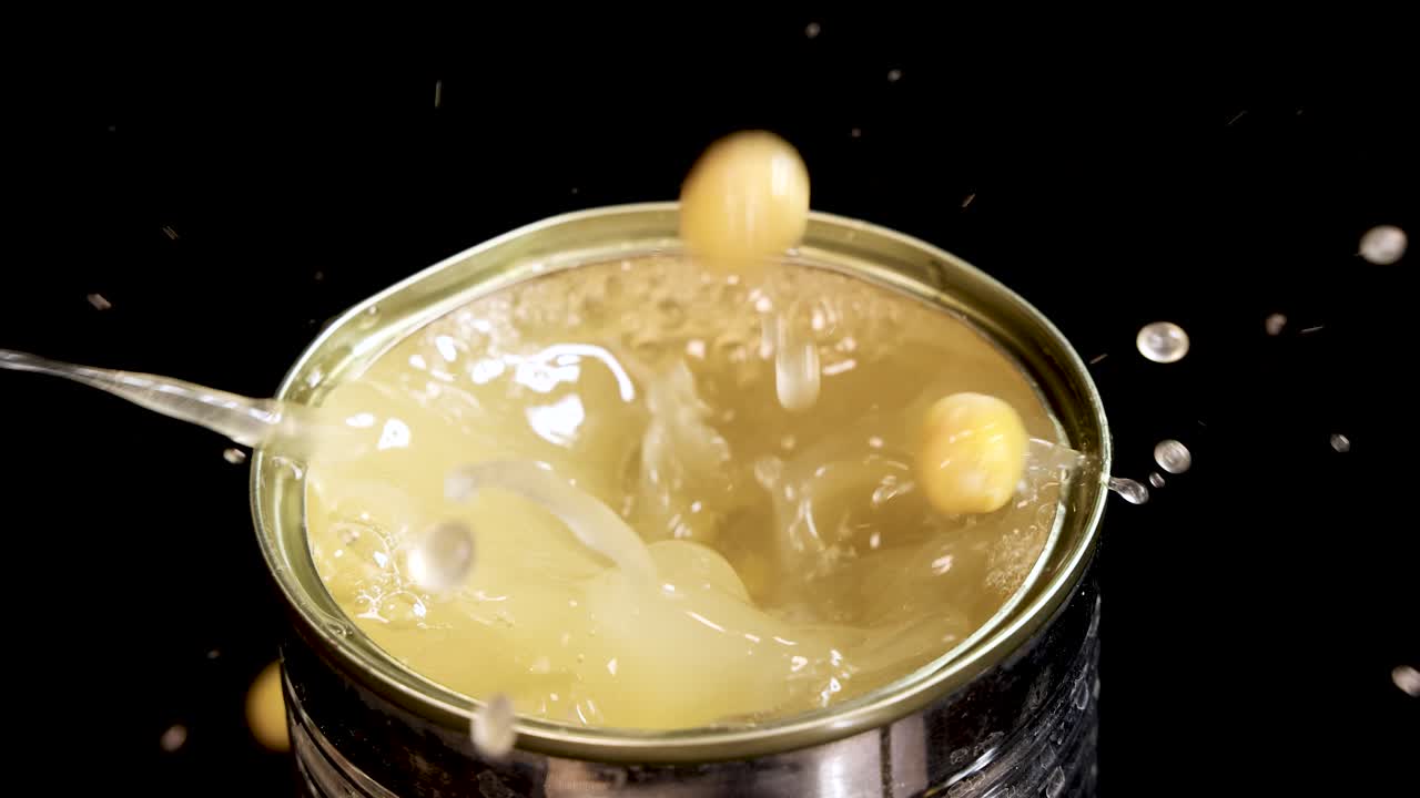 Multiple green peas fall into an open can filled with liquid, creating dynamic splashes. Studio lighting and black background highlight motion and texture