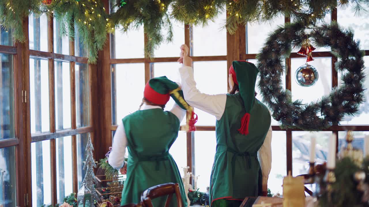 Joyful elves in green costumes are viewing the landscape from the window on the background of New Year decorations and Christmas wreath with a toy at day of New Year