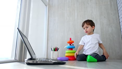 beautiful toddler in white t-shirt sits on the floor near toy pyramid. kid crawls to the laptop and lies in front of it.