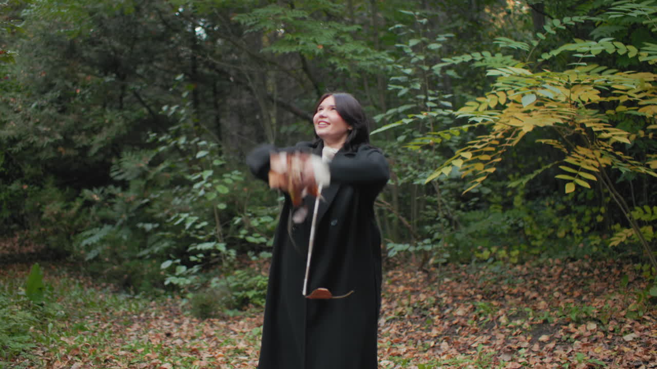 Nature lover tossing dry leaves upward while strolling through forest, smiling in long black coat and boots, golden branches around, playful carefree mood on brown leaf carpet in quiet park