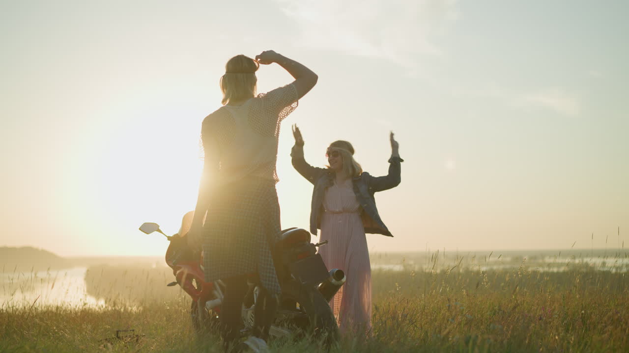 두 명의 여성, 머리 와 선글라스를 착용, 해가 지는 동안 오토바이 근처의 열린 잔디에서 기게 춤을 춘다