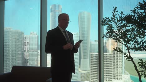 senior businessman silhouetted in a skyscraper office checks his smartphone and is surprised by a message in slow motion
