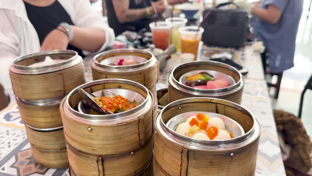 Hands reach for dim sum baskets on a sunlit table in a lively restaurant scene