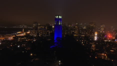 Colour-changing-Coit-memorial-tower-overlooking-downtown-San-Francisco-illuminated-skyline-during-4th-July-fireworks,-Aerial-orbiting-view