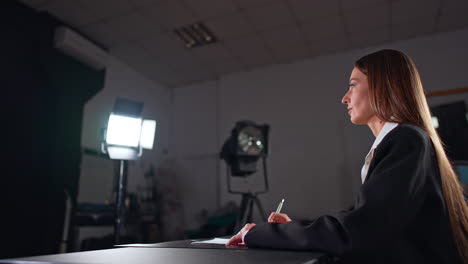 serious confident brunette caucasian woman in black jacket sits at desk. side view. studio lights are on ready for footage.