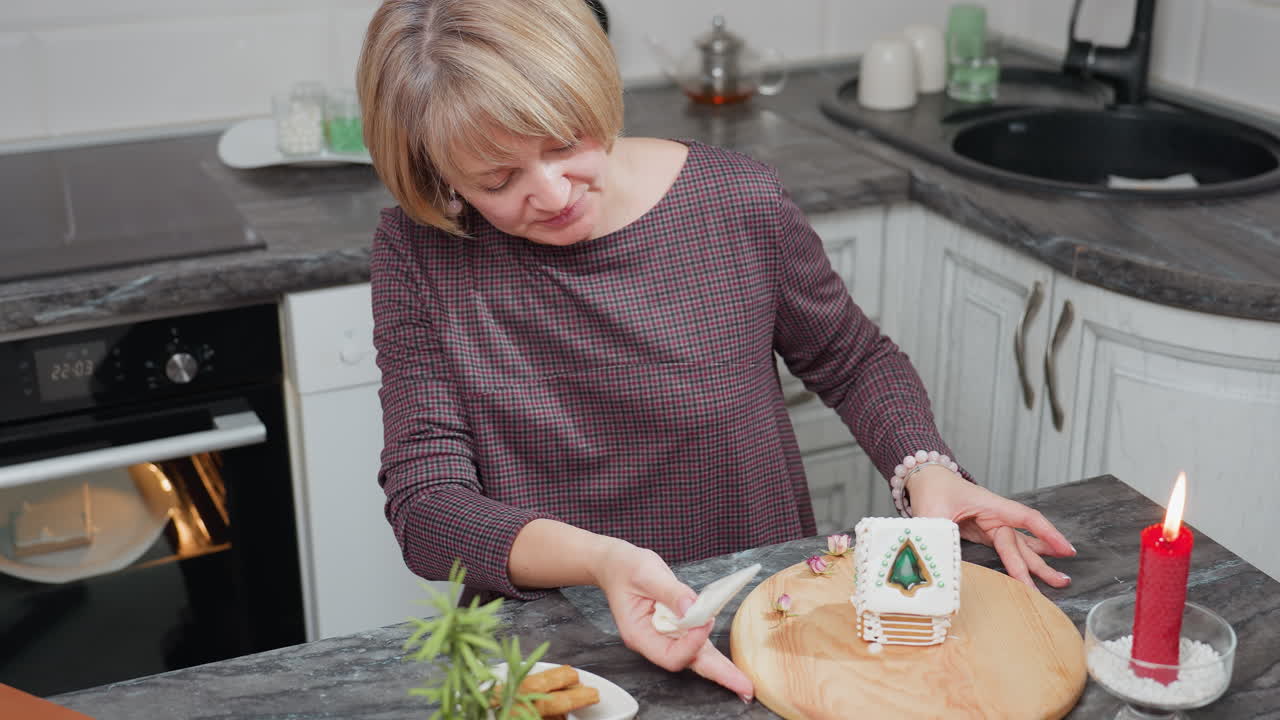 Kvinne dekorerer pepperkakehus forsiktig på et trebrett i et koselig kjøkken, kake stekes i ovnen og kjøkkenutstyr er synlig, en festlig scene med et rødt lys, cookies og planter i nærheten