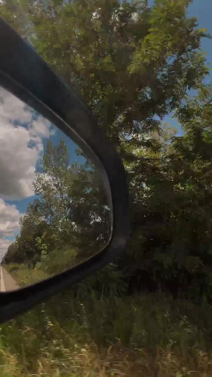 Car side mirror reflecting trees, farmland and a beautiful summer sky while driving along a country road. Road trip, freedom and rural travel concept. 4K vertical video.